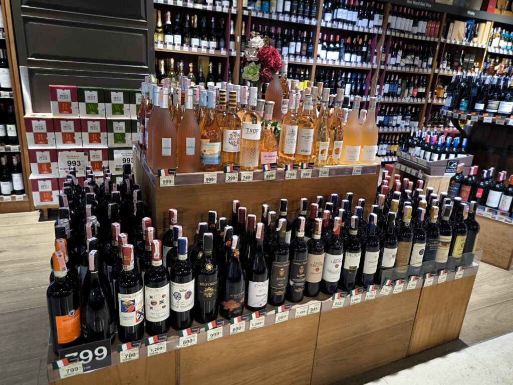 ไวน์ในร้านค้ามีไวน์หลากหลายชนิด เช่น ไวน์แดงและไวน์โรเซ่ ขวดไวน์หลายขวดวางเรียงกันอย่างเป็นระเบียบบนชั้นไม้พร้อมป้ายราคาที่มองเห็นได้ชัดเจน ขณะที่พื้นหลังเผยให้เห็นชั้นวางไวน์เพิ่มเติมที่เต็มไปด้วยไวน์หลากหลายชนิด.