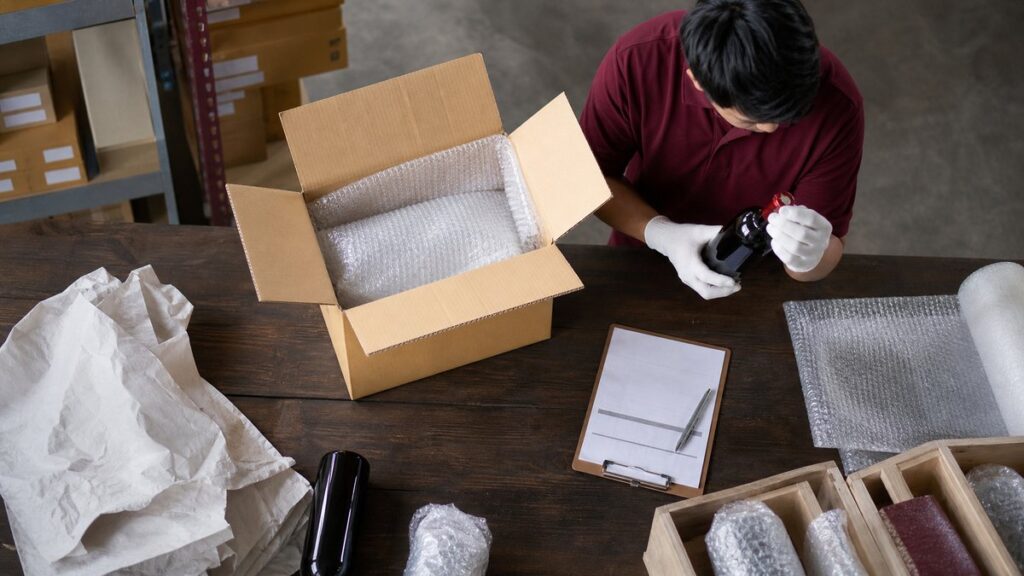 Overhead view of a careful packing process on a warehouse desk: an open shipping carton, bubble wrap, invoice papers, and a gloved hand checking a bottle’s neck seal. Cool neutral light falls evenly across the scene, while deep wine-red packaging accents and white padding suggest compliance, caution, and professional handling.